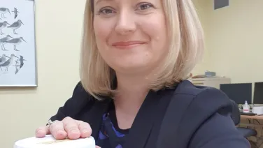 A woman in a business suit smiling at the camera while holding a jar containing preserved specimens, with illustrations of fish on the wall behind her.
