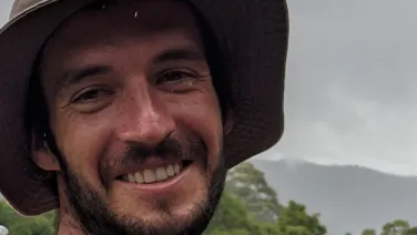 A smiling man wearing a wide-brimmed hat stands outdoors with a cloudy mountain backdrop.