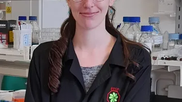 A woman with glasses and a ponytail, wearing a lab coat, smiles in a laboratory setting.