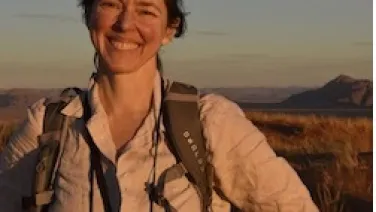 A woman smiling in a field at sunset, wearing a backpack and a camera around her neck.