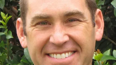 A close-up portrait of a smiling man with short brown hair standing in front of a leafy green background.