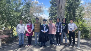 A group of nine people poses for a photo outdoors, with one person wearing graduation attire.