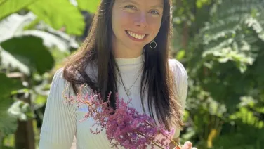 A woman smiling, holding pink flowers while standing in a lush garden.
