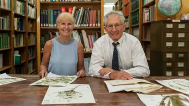 An older man and woman smiling at the camera while seated at a library table with books, maps, and botanical prints.