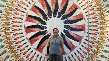 A woman smiling in front of a colorful wall display featuring a radiating pattern of feather illustrations.