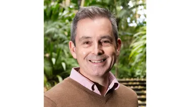A smiling middle-aged man wearing a brown sweater over a pink shirt, standing in front of lush greenery.