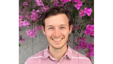 A smiling man in a pink striped shirt standing in front of a wall covered with pink bougainvillea flowers.