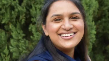 A woman with long dark hair smiling outdoors with green foliage in the background.