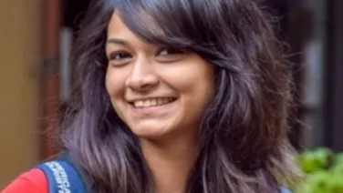 A young woman with shoulder-length dark hair smiles broadly, wearing a red top and carrying a blue backpack.