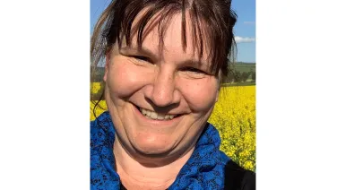 A smiling woman with sunglasses on her head standing in front of a vibrant yellow rapeseed field under a clear blue sky.