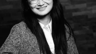 A black and white portrait of a smiling woman wearing glasses, seated with one hand resting on an acoustic guitar, against a brick wall background.