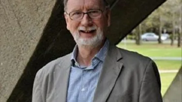 An older man with a beard and glasses, smiling and standing under an archway.