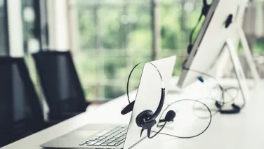 A headset hangs on a laptop in a bright office with windows in the background.