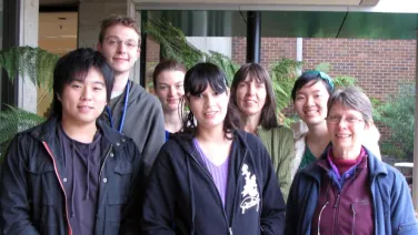 A group of seven diverse people smiling and standing together in front of a building with greenery.