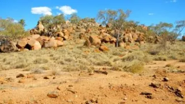 A dry, sparse landscape featuring red rocky outcrops and scattered scrub under a clear blue sky.